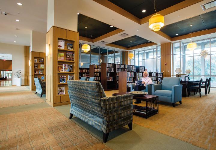 A NewBridge on the Charles resident sits and reads a magazine in a contemporary library with ample seating and open shelving for books and periodicals. 