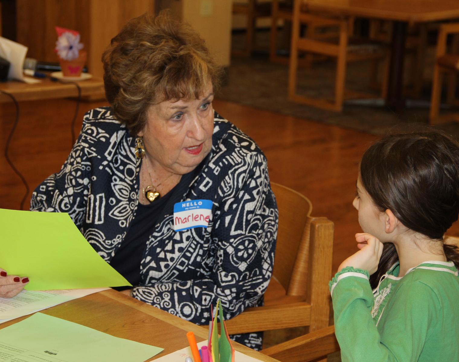 An older woman is seated at a table with a young girl standing next to her. They are both smiling and looking at the camera. A craft project sits in front of them.