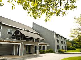 Three-story gray shingled apartment building, with a driveway in front of the main entrance. Trees and grass surround the building.