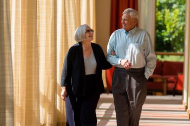 An older woman links her arm through an older man's arm as they walk down a hallway smiling.