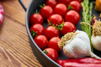 Skillet with bright red grape tomatoes, unpeeled onions and garlic, and red pepper on wooden board with red and white checkered fabric.