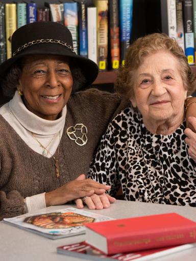 Two older women sit together in a library at a table with books and magazines.