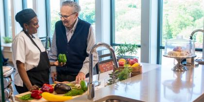 A male resident stands and talks with a female cook while she is holding a green bell pepper.