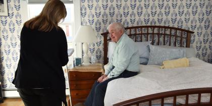 A Home Health worker stands and talks with a male patient while he sits in his bed.