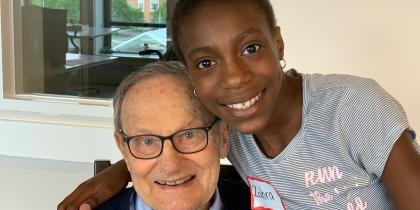 An elder male embraces a child volunteer in a hug as they both smile