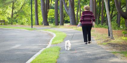 An older adult woman walks on a trail with her dog's leash in one hand and a walking stick in the other.
