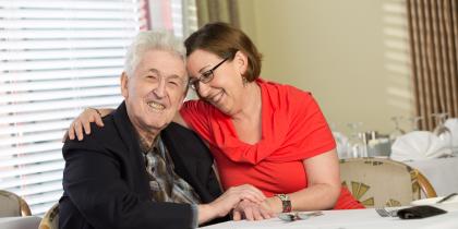 A male senior living resident and his adult daughter embrace in a hug at a dining table.