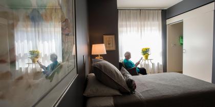 An older adult woman sits in a chair by the window in her room and reads