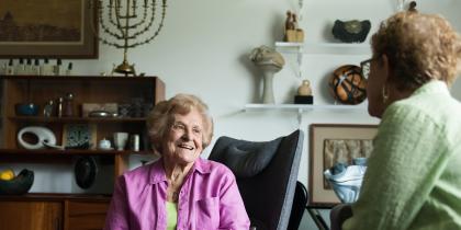 An older woman sitting in her room sits and talks with another woman.