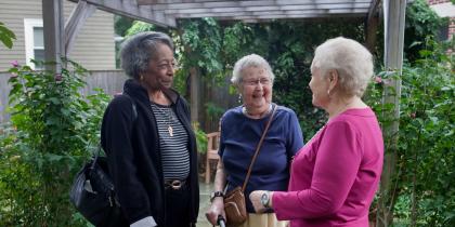 Three women stand together smiling outside surrounded by greenery 