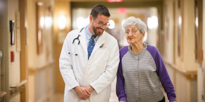 A female patient using a walker walks down a hallway smiling and talking with a male doctor 