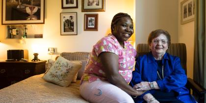 A female caregiver sits on a bed while a female patient sits on a chair next to the bed