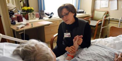 Rabbi Sara Paasche-Orlow  sits in a chair and holds a patients hand while talking to her 