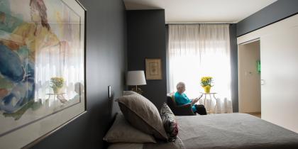 An older adult woman sits in a chair by the window in her room and reads