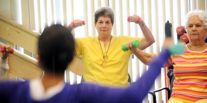 Two senior women lift small weights, following the instruction of their class leader.