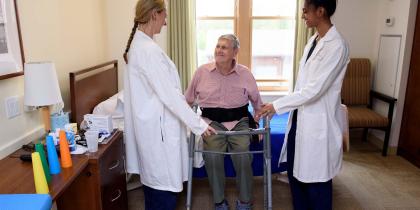 Older man sitting on bed smiling up at two nurses who are readying a walker