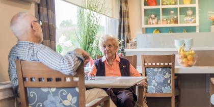 Sitting in a dining area, an older woman in a pink shirt and purple vest talks with a man in a black and white plaid shirt whose back is to the camera.