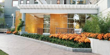 Entryway of Goldman Residences at 1550 Beacon Place, Brookline, MA- Granite textured building with two-tiered white awning, orange tile, benches and orange flowers in raised beds.