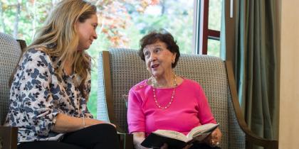 Older woman in pink shirt with a book open on her lap, speaking with a woman.