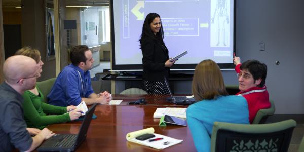 A female scientist from the Marcus Institute stands in front of a screen with a slide that has an image of a skeleton and says "Protein & Osteoporosis;" she is standing in front of a table where four other scientists sit