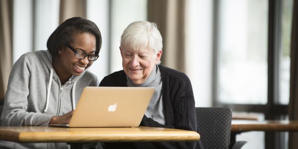 A young woman and older woman sit at a table looking at a lapto; they are smiling