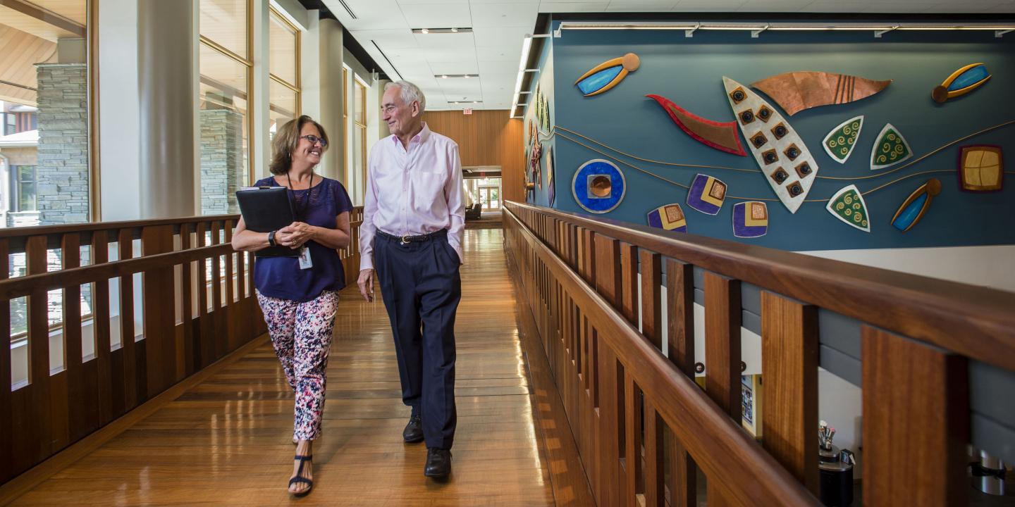 An older man in an oxford shirt and navy trousers walks across the community center bridge with the NewBridge on the Charles move-in coordinator.