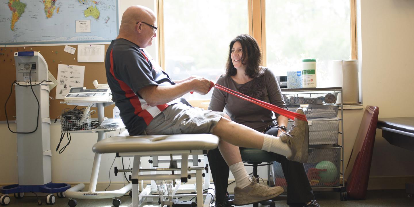 In a sunny gym room, a physical therapist encourages at a man using a red elastic band to do a leg exercise.