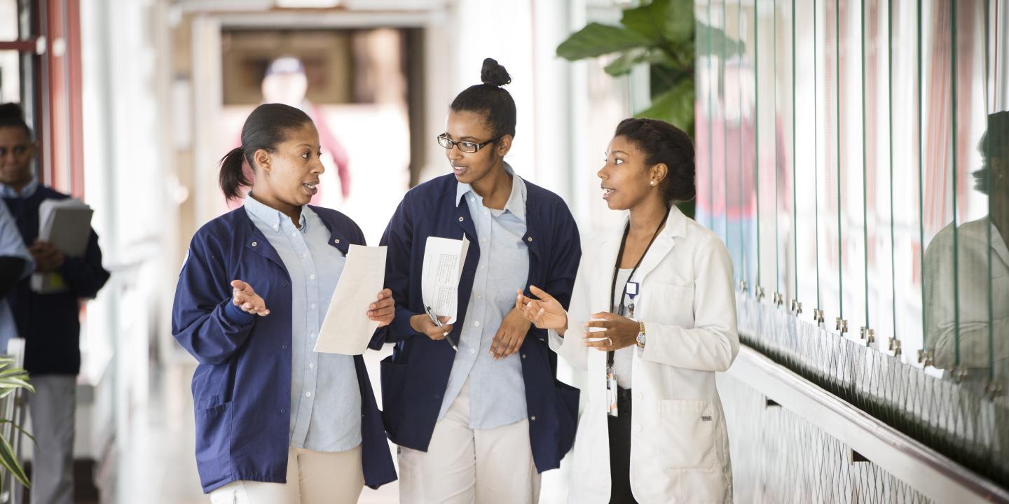 Two female student nurses and a female nurse stand talking in a hallway at Hebrew Rehabilitation Center in Boston. The students are wearing short blue coats and the nurse is wearing a white coat.