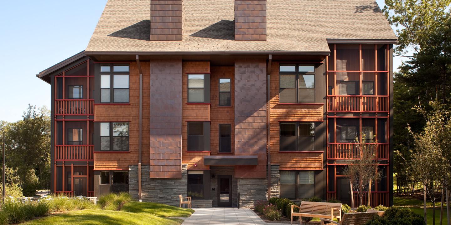 Villa exterior showing warm terra cotta tones on the building façade, a communal courtyard with outdoor seating, and greenery all around.