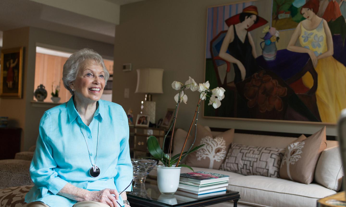 Attractive smiling older woman in a turquoise shirt sits on a couch in her well-appointed independent living apartment.