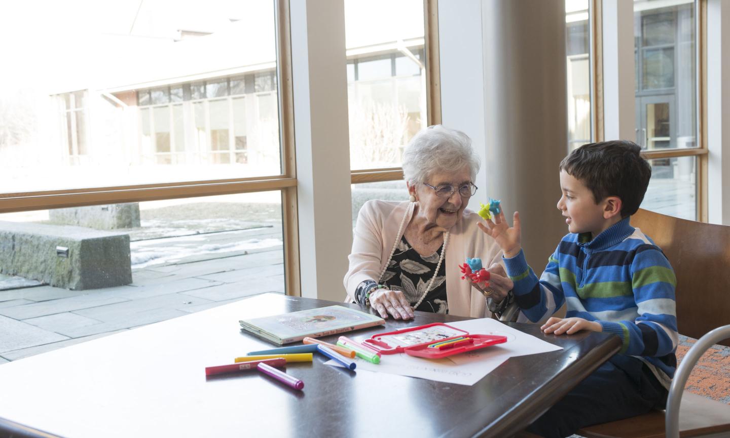 Older woman sits at a table with a young boy as they play with colorful finger puppets.