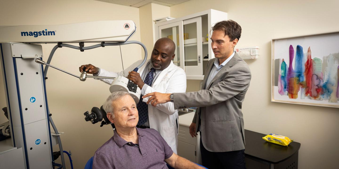 An elderly patient receives TMS treatment for depression at the Wolk Center for Memory Health in Boston. He is seated with a calm expression on his face. A technician holds an electromagnetic coil next to his head while the neuropsychologist looks on.