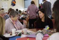 An older woman at a middle-aged woman sit talking to each other at a table covered with construction paper, craft supplies, and coffee cups.