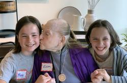 An older woman kisses the cheek of a young girl, with another young girl holding the woman's hand and smiling.