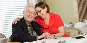 A male senior living resident and his adult daughter embrace in a hug at a dining table.