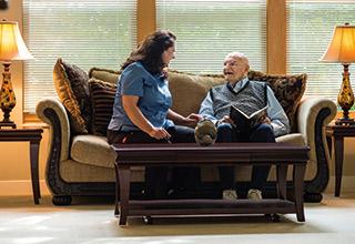 A young woman and an elder man sit on a couch. The man is holding a book and looking up and smiling at the woman.