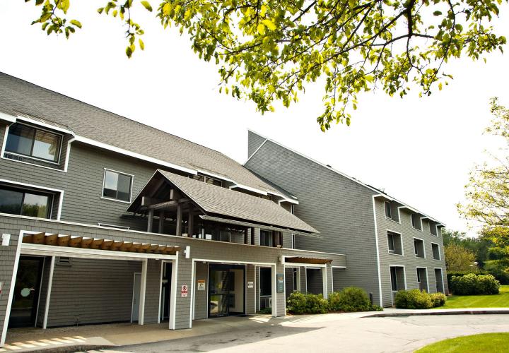 Three-story gray shingled apartment building, with a driveway in front of the main entrance. Trees and grass surround the building.