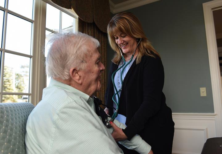 Woman with stethoscope takes blood pressure of man seated in arm chair.