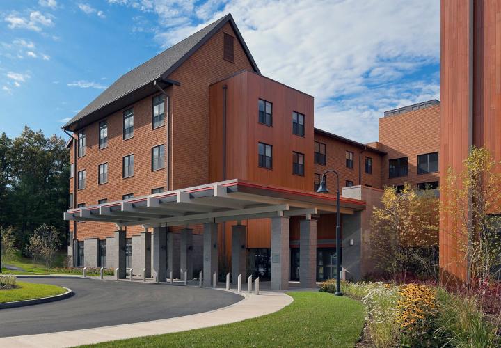 A curved entrance road leads to the Health Care Center at NewBridge on the Charles, home to Hebrew Rehabilitation Center Dedham. The golden brown exterior of the building matches the color of the early autum leaves on the surrounding trees.