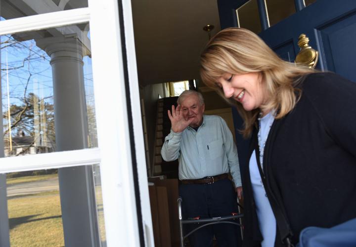 At the doorstep of his house, a nurse from Hebrew SeniorLife Home Health greets older man using a walker